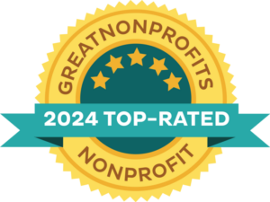 This image shows the Great Nonprofits 2024 Top-Rated badge. It is a golden yellow circle with serated edges. In the center is a smaller green circle with five gold stars in an arc at the top of the circle. Across the lower third is a green banner that says, "2024 Top-Rated"