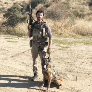 This photo shows Dita and her human handler. Dita is a Belgian Malinois dog sitting on a flat dirt road and wearing a military harness. The handler is standing next to her, dressed in khaki pants and tactical vest, holding her leash in one hand and a rifle pointing skyward in his other hand. In the background of the photo are dried grasses and small shrubs, suggesting a semi-arid climate.