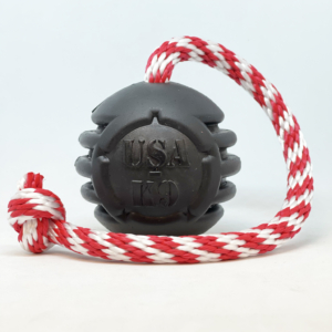 This photo shows a sturdy black rubber ball with grooves in the sides, attached to a red and white striped rope. This sturdy dog toy is called, "USA-K9 Magnum Black Stars and Stripes Ultra-Durable Rubber Chew Toy, Reward Toy, Tug Toy, and Retrieving Toy." The manufacturer is SodaPup in Colorado. Words molded into the black rubber say, "USA-K9," which is one of SodaPup's product lines. Dita's Final Donation Drop 2024 Toy Drive is featuring this toy.